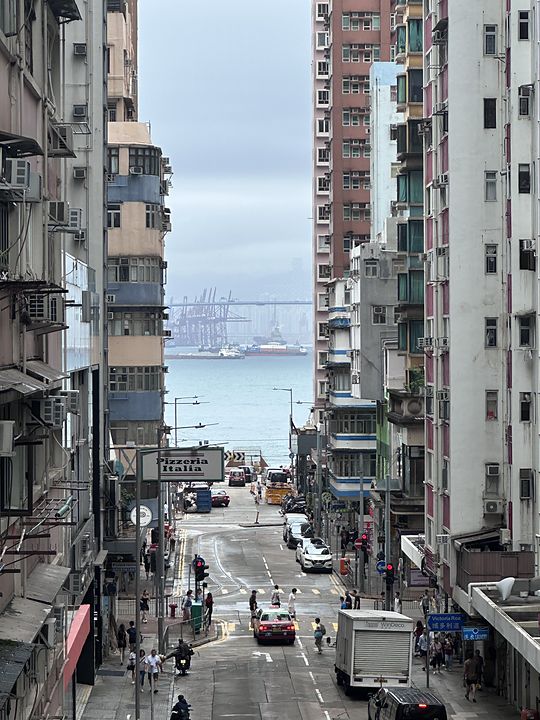 A steep Hong Kong street running down to the harbour, container cranes on the far shore