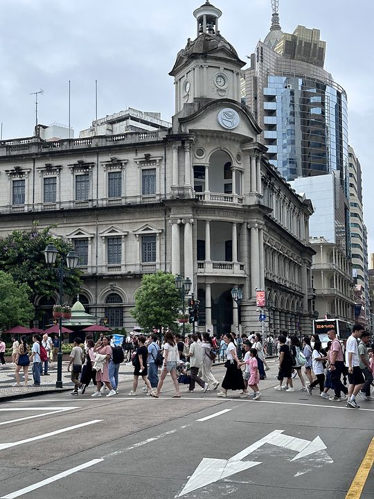 A Portuguese colonial building with a clock tower and a lotus emblem in central Macau, crowds crossing in front