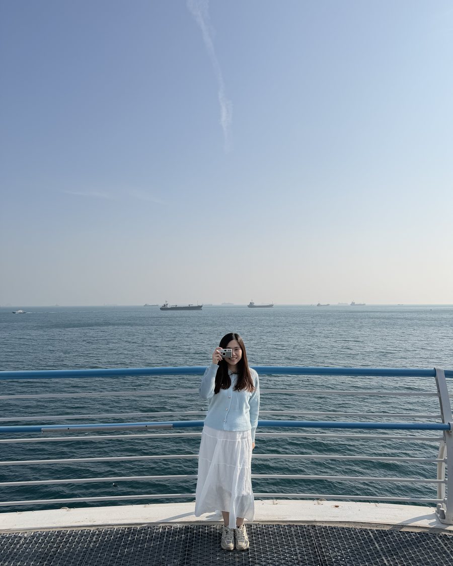 Ivy holding a compact camera at the sea railing on Songdo in Busan, cargo ships anchored along the horizon