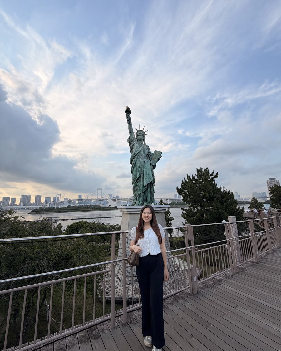 Ivy on the Odaiba waterfront boardwalk in Tokyo, below the replica Statue of Liberty, with Rainbow Bridge across the bay behind her
